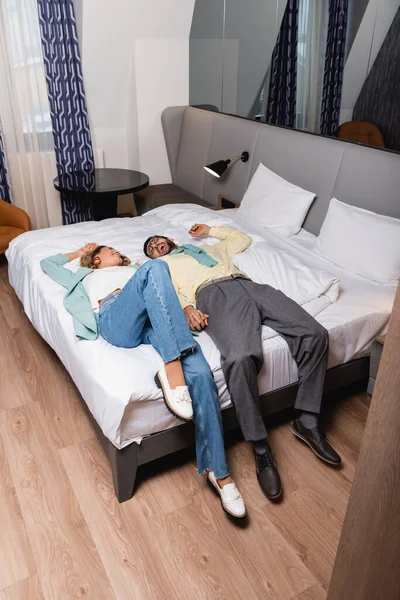 Alegre pareja interracial cogido de la mano mientras está acostado en la cama del hotel - foto de stock