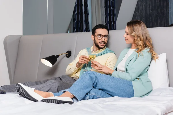 Sonriente pareja interracial brindis con vino en la cama del hotel - foto de stock