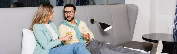 Pareja interracial sosteniendo copas de vino en la cama en la habitación del hotel, pancarta - foto de stock