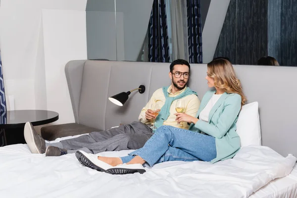 Hombre musulmán con copa de vino mirando a su novia en la cama del hotel - foto de stock