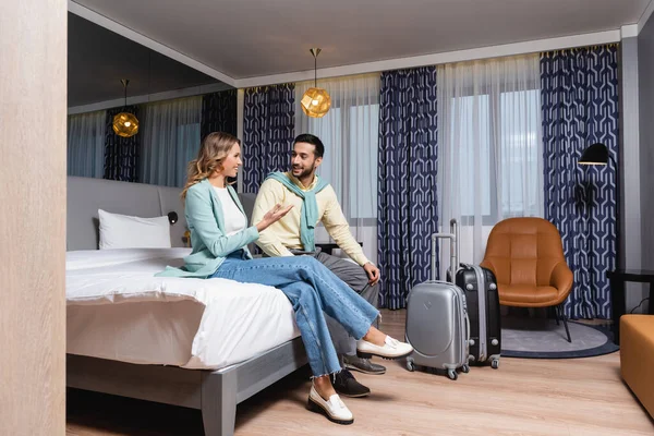 Smiling woman pointing with hand near suitcases and muslim boyfriend on bed in hotel — Stock Photo