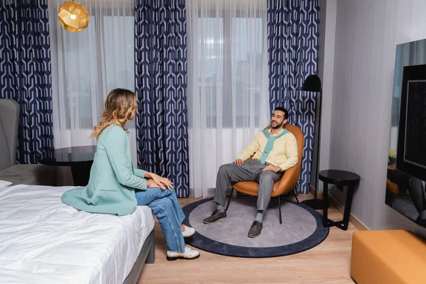 Hombre árabe sonriente sentado en un sillón cerca de su novia en la cama en el hotel - foto de stock