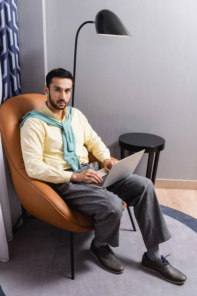 Hombre árabe mirando a la cámara mientras usa el ordenador portátil en el hotel - foto de stock