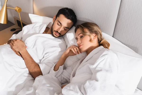 Vista de ángulo alto de pareja interracial en albornoces durmiendo en la habitación del hotel - foto de stock