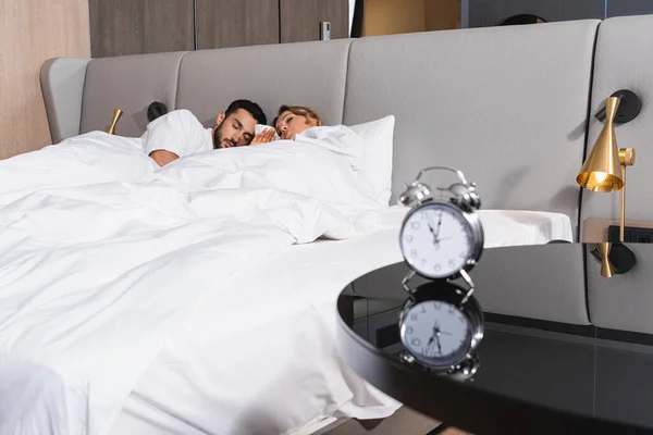 Pareja interracial durmiendo en ropa de cama blanca cerca del despertador en la mesa en primer plano borroso en el hotel - foto de stock