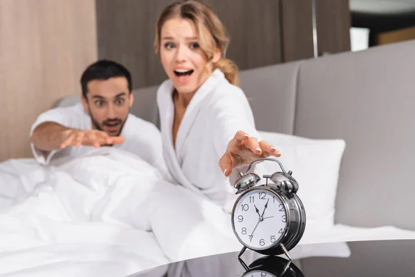Alarm clock near excited woman and muslim man on hotel bed on blurred background — Stock Photo