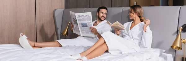 Sonriente pareja interracial con periódico y libro sentado en la cama del hotel, pancarta - foto de stock
