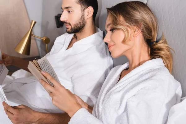 Mujer joven leyendo libro cerca de novio musulmán con noticias sobre fondo borroso en el hotel - foto de stock