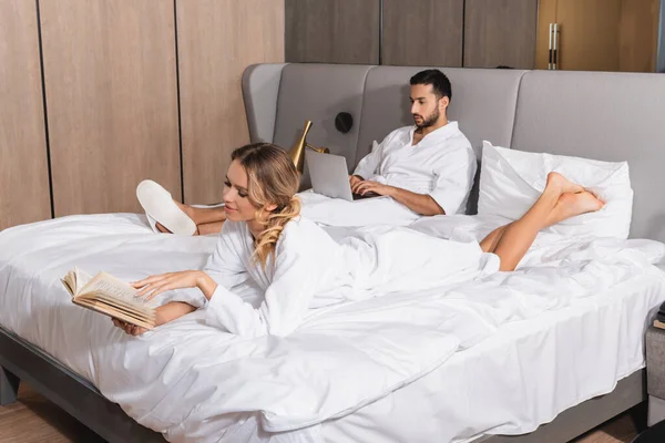 Mujer con libro cerca novio árabe con portátil en la cama en la habitación de hotel - foto de stock