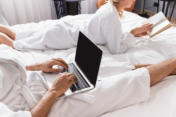 Обрезанный вид человека, использующего ноутбук с экраном-бланком рядом с подругой с книгой на кровати отеля — стоковое фото