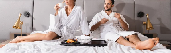 Muslim man holding coffee cup near girlfriend and breakfast on hotel bed, banner — Stock Photo