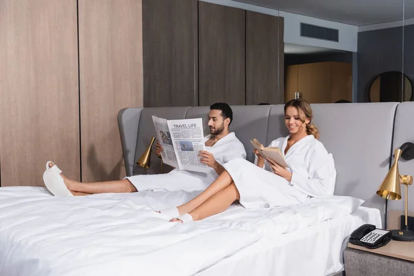 Smiling woman reading book near arabian boyfriend with newspaper in hotel — Stock Photo