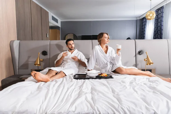 Young woman holding cup near breakfast and muslim boyfriend on hotel bed — Stock Photo