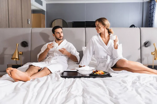 Mujer sonriente con taza mirando al hombre árabe cerca del desayuno en la cama en el hotel - foto de stock