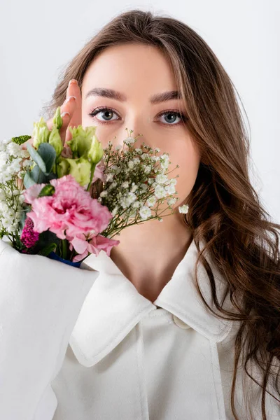 Mujer morena mirando a la cámara cerca de flores en manga de abrigo aislado en gris - foto de stock