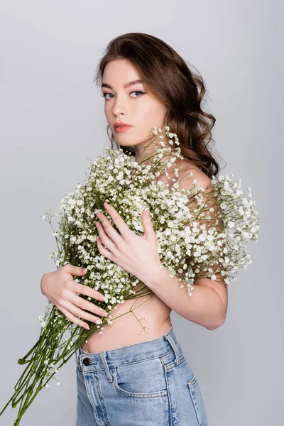 Donna bruna senza maglietta in jeans con mazzo di piccoli fiori bianchi isolati su grigio — Foto stock