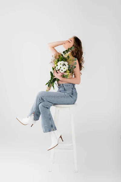 Donna senza maglietta in jeans che tiene fiori sulla sedia isolata su grigio — Foto stock