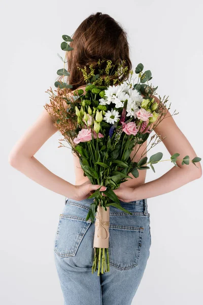 Vista posterior de una joven en jeans sosteniendo diferentes flores aisladas en gris - foto de stock