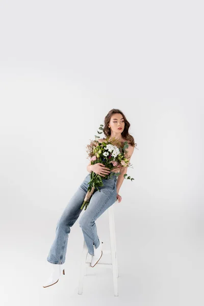 Shirtless model in shoes and jeans holding bouquet on chair on grey background — Stock Photo