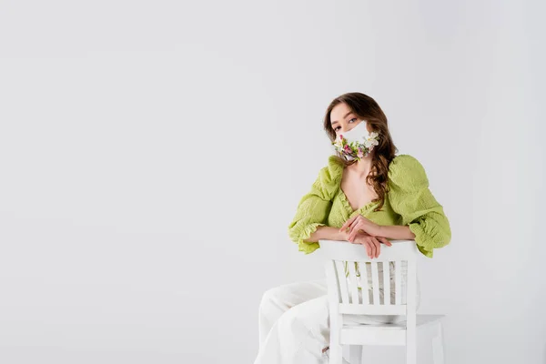 Modèle Brunette avec fleurs sur masque médical regardant caméra sur chaise isolée sur gris — Photo de stock