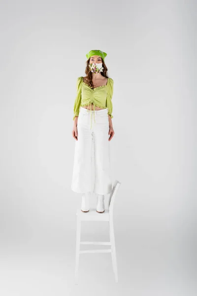 Donna alla moda in maschera medica con fiori e velo in piedi sulla sedia isolata su grigio — Foto stock