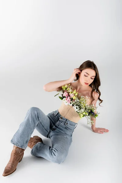 Брюнетка в блузке с цветами и джинсами сидит на сером фоне — стоковое фото