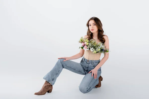 Mujer morena en zapatos marrones, jeans y flores en blusa sobre fondo gris - foto de stock