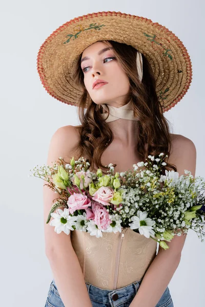 Кучерява модель в сонячному капелюсі з квітами в блузці дивиться ізольовано на сірому — стокове фото