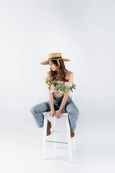 Trendy woman in straw hat and floral bouquet in blouse posing on chair on grey background — Stock Photo