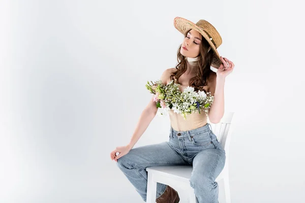 Young woman with flowers and sun hat sitting on white chair isolated on grey — Stock Photo