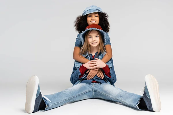 Happy african american girl embracing cheerful, trendy friend sitting on grey — Stock Photo