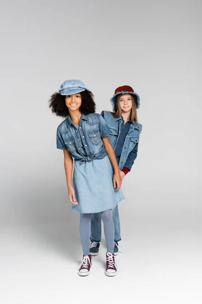 Ansicht von stilvollen interrassischen Mädchen in trendigen Kleidern und Gummischuhen, die auf grau posieren — Stockfoto