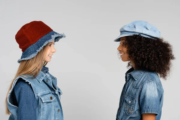 Vista lateral de duas meninas inter-raciais em roupas da moda e chapéus sorrindo isolado no cinza — Fotografia de Stock