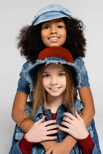 Happy interracial kids in fashionable clothes and hats embracing and looking at camera isolated on grey — Stock Photo