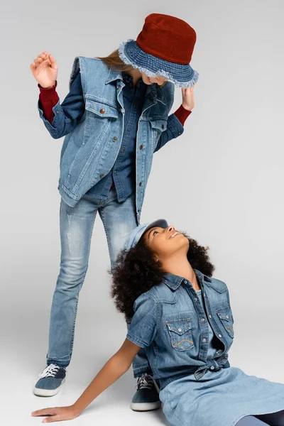 Модна дівчина в стильному джинсовому одязі стоїть над щасливим афроамериканським другом, сидячи на сірому — стокове фото