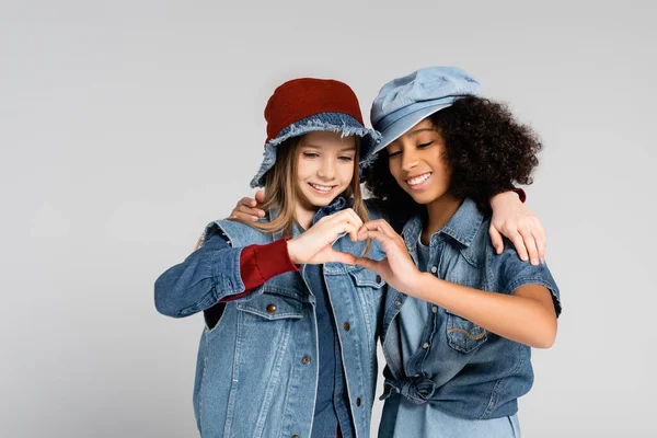 Dos amigos interracial alegres y de moda abrazando y mostrando el signo del corazón con las manos aisladas en gris - foto de stock