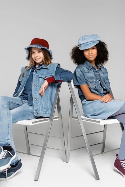Fashionable interracial girls sitting on chairs and smiling at camera on grey — Stock Photo