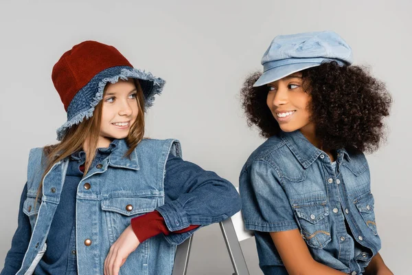 Cheerful interracial friends in trendy denim clothes looking at each other isolated on grey — Stock Photo