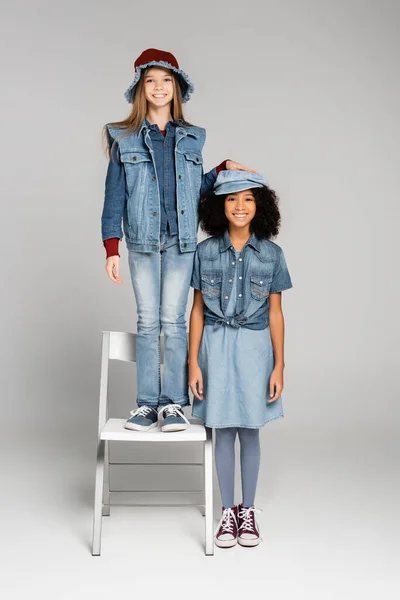 Веселая модная девушка, стоящая на стуле рядом с африканским американским другом в джинсовой одежде на сером — стоковое фото
