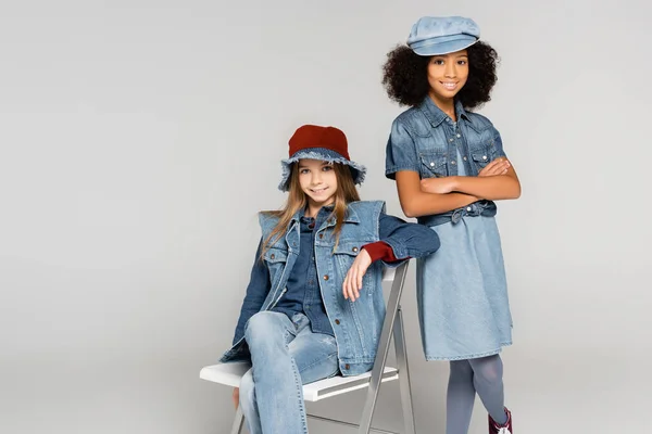 Fashionable african american girl standing with crossed arms near friend sitting on chair on grey — Stock Photo