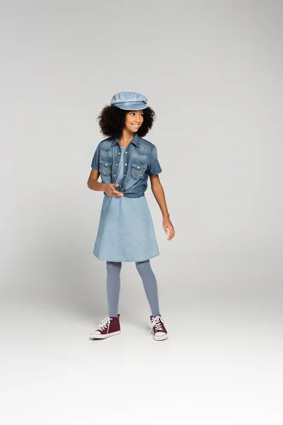 Fashionable african american girl in denim clothes, cap and gumshoes posing on grey — Stock Photo
