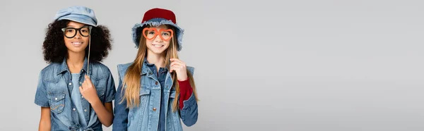 Веселые межрасовые девушки в модной джинсовой одежде, с вырезанными из бумаги очками, изолированными на сером, баннер — стоковое фото