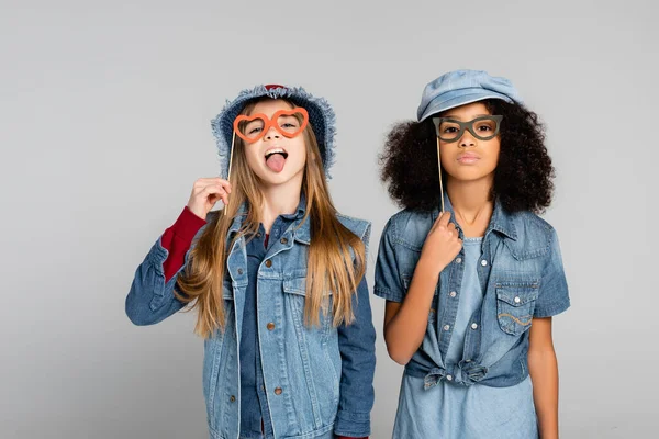 Trendiges Mädchen, das die Zunge herausstreckt, während es mit einem afrikanisch-amerikanischen Freund eine Brille auf grau hält — Stockfoto