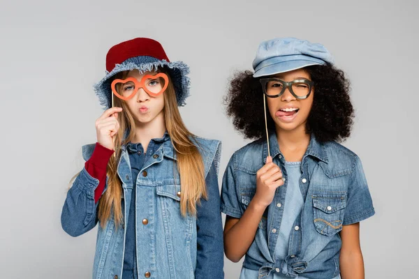 Cheerful, stylish interracial girls holding paper cut spectacles and grimacing isolated on grey — Stock Photo