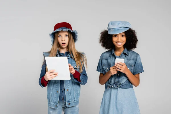 Niños interracial emocionados en ropa de moda utilizando teléfono inteligente y tableta digital aislado en gris - foto de stock