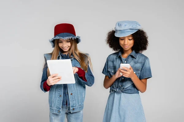 Smiling multicultural girls in denim clothes and hats using gadgets isolated on grey — Stock Photo