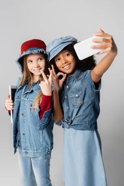 Fashionable interracial girls showing okay and victory signs while taking selfie on grey — Stock Photo