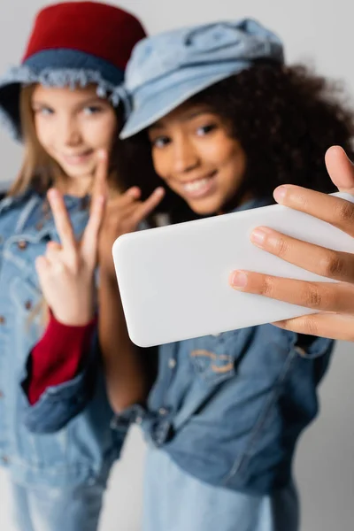Meninas inter-raciais felizes mostrando sinais de vitória e rock ao tomar selfie no fundo cinza e borrado — Fotografia de Stock
