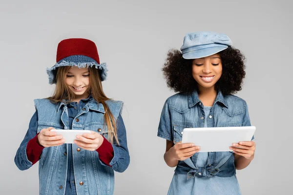 Joyful interracial kids in fashionable denim clothes and hats using smartphone and digital tablet isolated on grey — Stock Photo