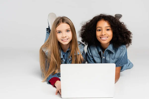 Complacido, chicas interracial de moda sonriendo a la cámara mientras se encuentra cerca de la computadora portátil en gris — Stock Photo